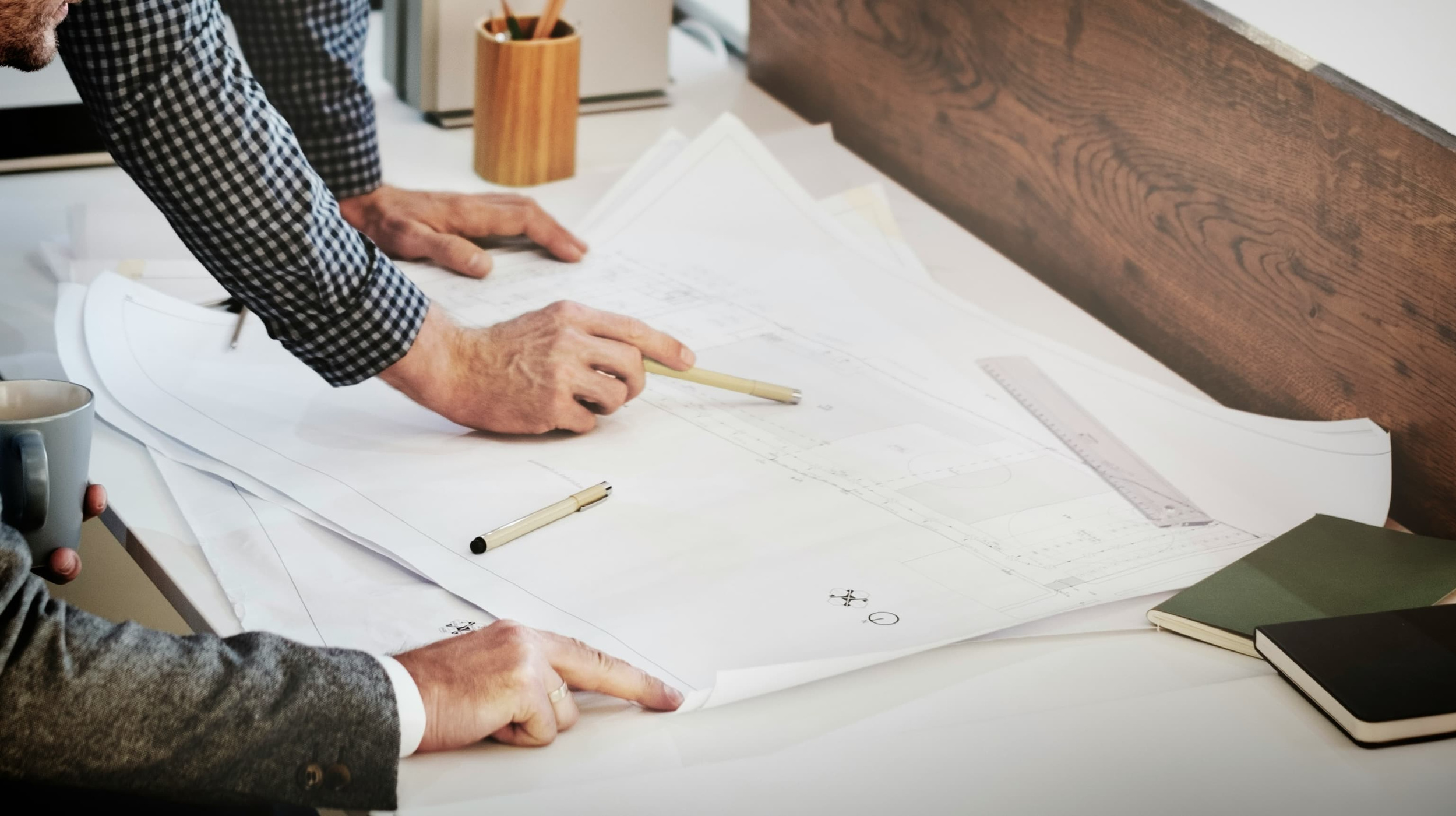 Twee personen bespreken technische bouwplannen op een witte bureau; de een wijst met vinger naar de tekening, de ander houdt een pen en mok vast, liniaal en notitieblokken op tafel.