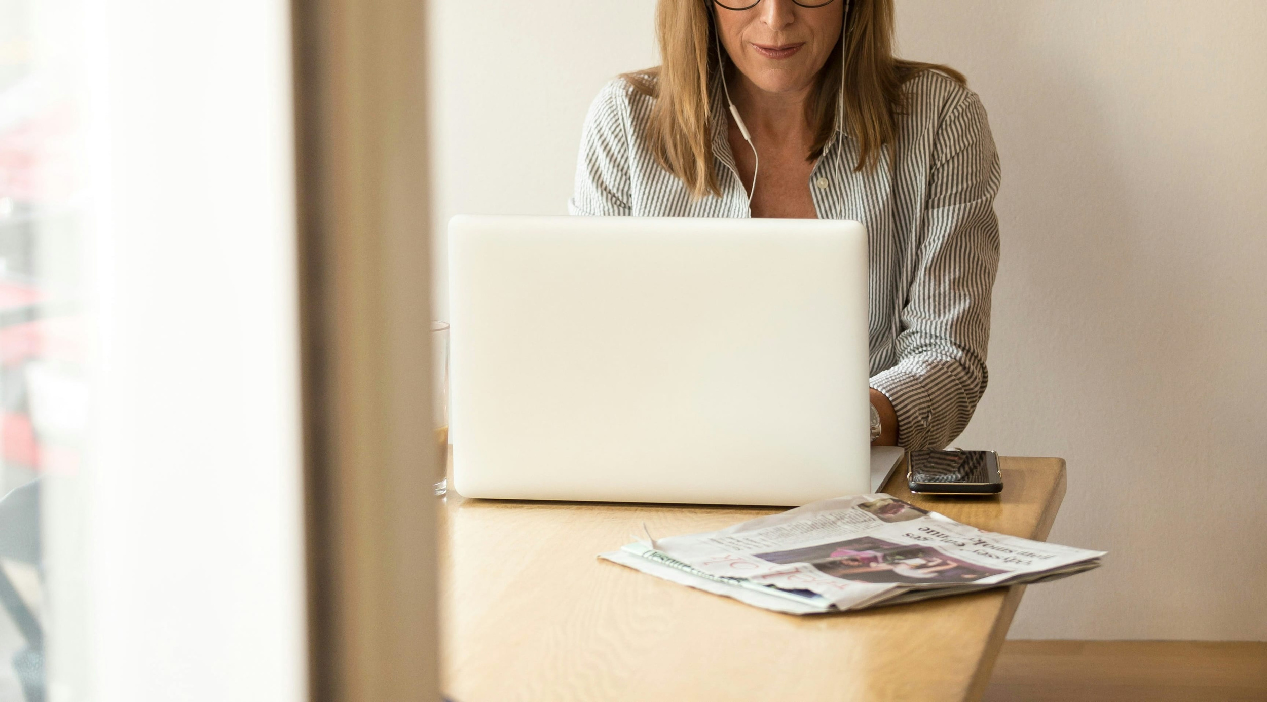 Persoon met bril en gestreept shirt werkt geconcentreerd aan een laptop aan tafel, met smartphone en krant; witte oortjes en glas op tafel, lichte binnenruimte.