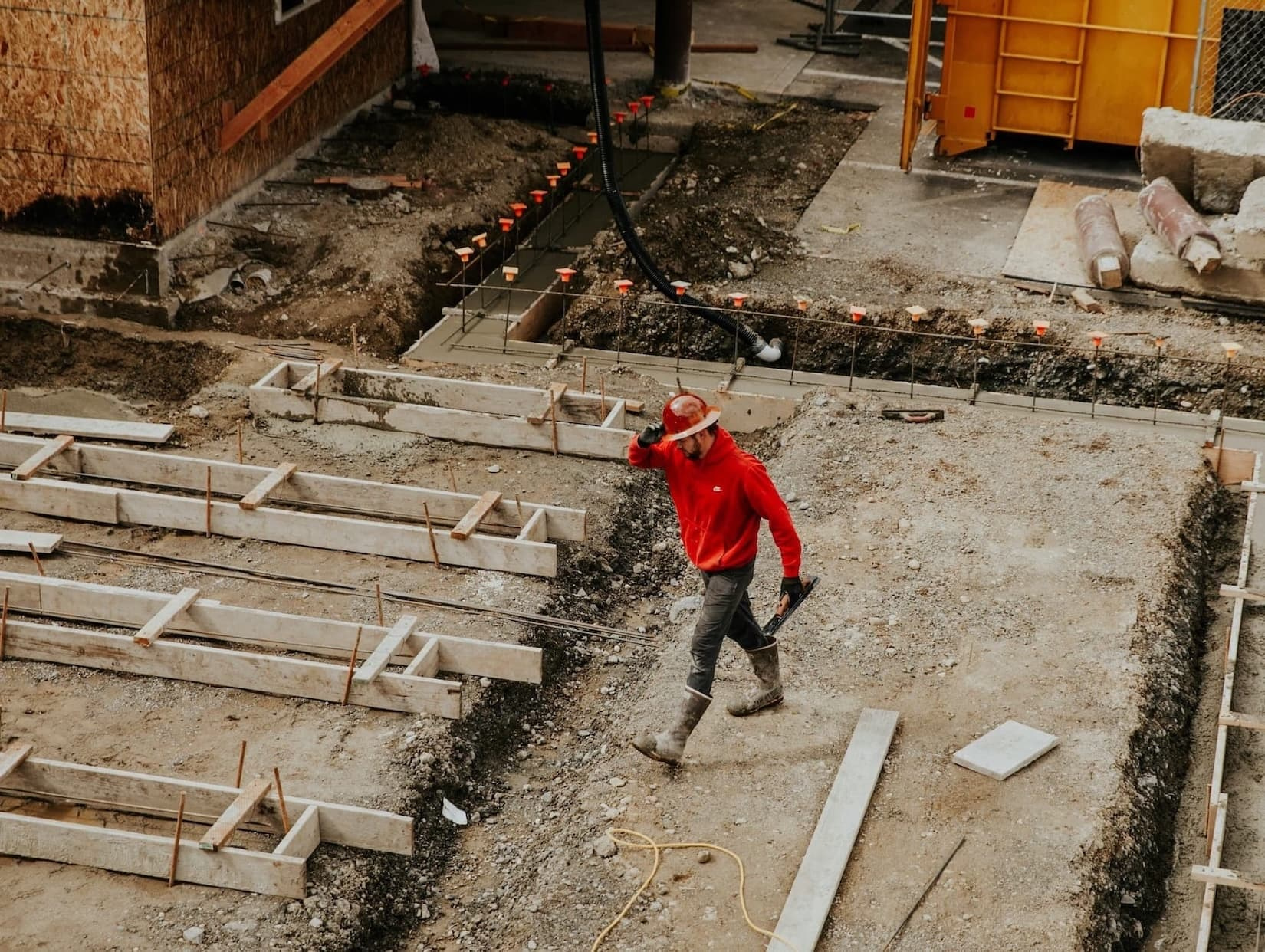 Bouwvaker in rode hoodie en helm loopt over een werf; op de voorgrond houten bekisting en verse betonfundering met wapening, op de achtergrond bouw en gele constructie.